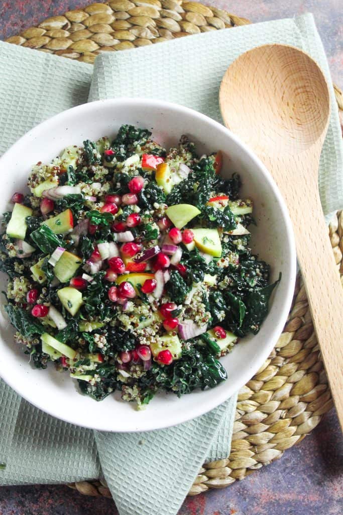 Bowl of kale and pomegranate salad with a lemon thyme vinaigrette. Shown in a white bowl on a woven mat with a wooden serving spoon. 