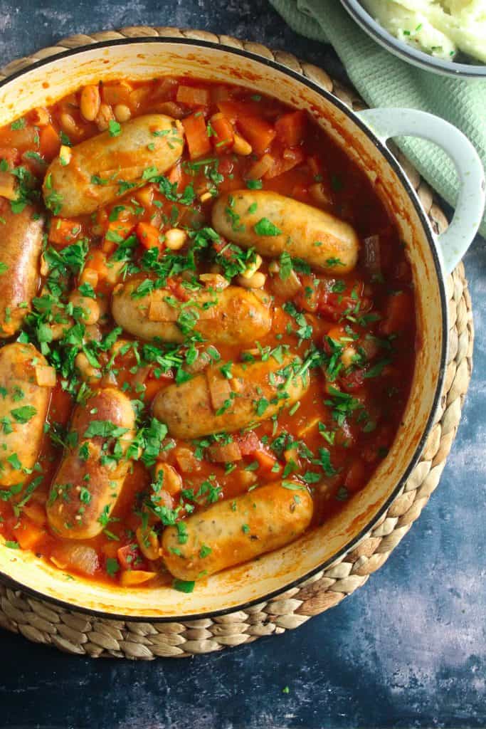 Fat pork sausages in a red casserole sauce with cannellini beans and carrots. Garnished with fresh chopped parsley in a blue casserole dish on a wicker place mat. 