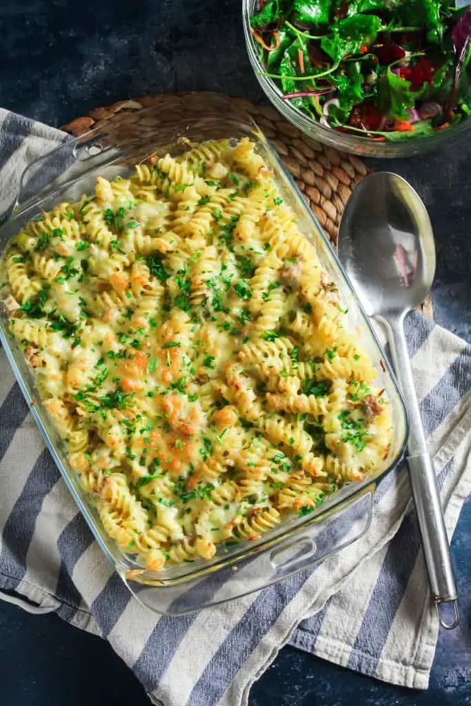 A baking dish full of fusilli pasta in a creamy tuna sauce. Topped with grilled cheddar cheese and chopped parsley. 