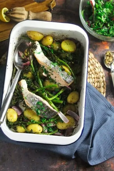 White enamel roasting dish containing two baked salmon fillets resting on sliced, roast baby potatoes, tenderstem broccoli and red onion.