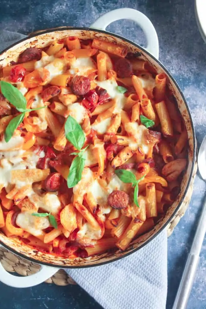 Rigatoni pasta with chunks of chorizo sausage, chicken pieces, bell pepper and tomato, topped with melted cheese and baked in a large blue casserole dish.