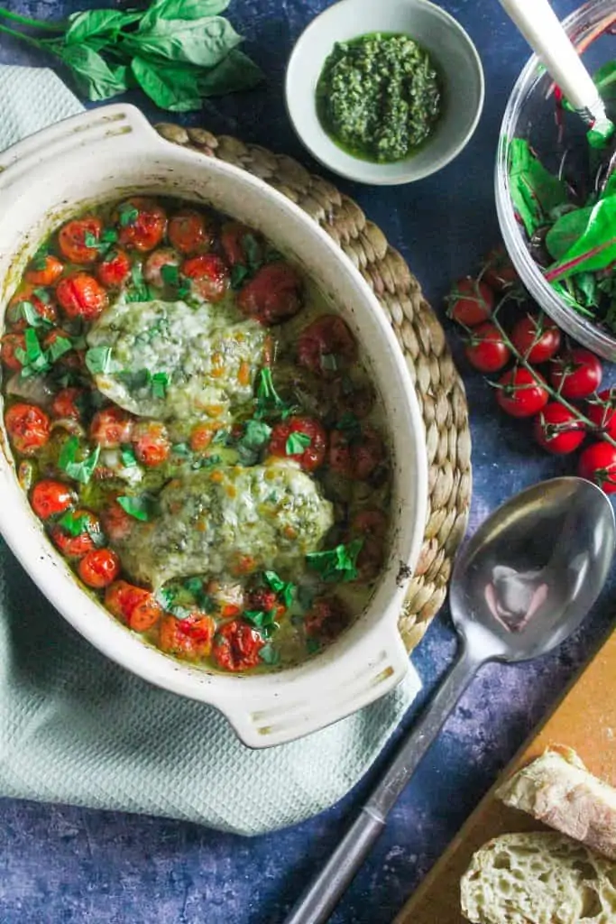 Oval casserole dish containing chicken breasts covered in pesto and melted mozzarella, surrounded by burst cherry tomatoes. 