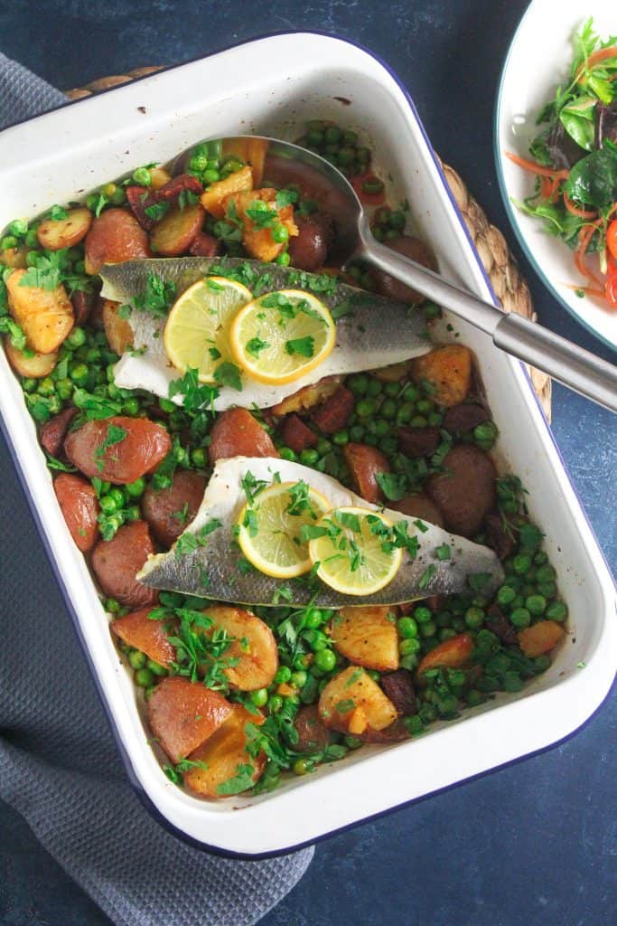 Sea bass fillets garnished with lemon slices. In a roasting tin on a bed of peas, sliced red-skinned baby potatoes and chunks of chorizo sausage.