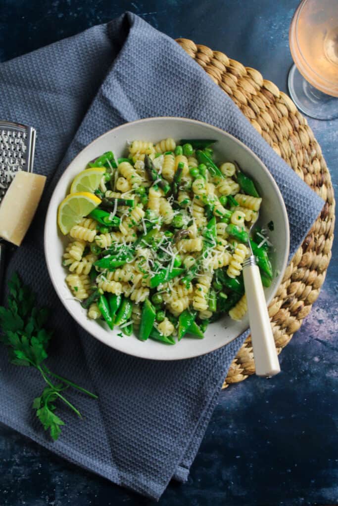 Pasta Primavera with Peas and Asparagus in a white pasta bowl sitting on a blue cloth with a wedge or parmesan next to it. 