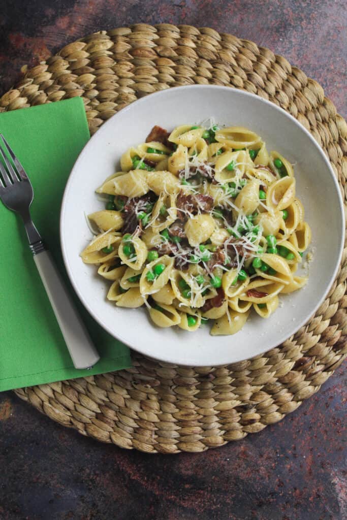 White bowl on woven mat next to a green napkin. The bowl contains pasta shells with diced bacon and peas, topped with grated cheese. 