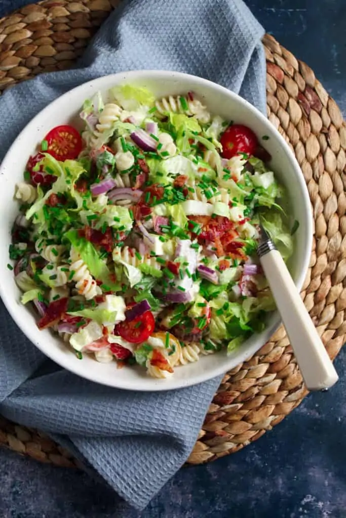 White bowl on a woven mat containing BLT Pasta Salad - fusilli pasta, crispy bacon pieces, shredded lettuce, cherry tomatoes, sliced red onion and mozzarella topped with fresh cut chives. 