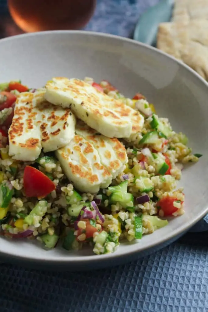 Close up of a bowl of bulgur wheat salad topped with slices of thick cut fried halloumi cheese. 