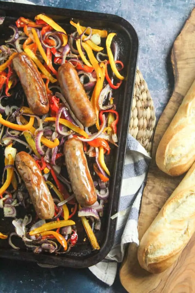 Baking tray loaded with crisp cooked pork sausages and roasted sliced onions and peppers. The tray is lying next to a board with two baguettes lying on it.