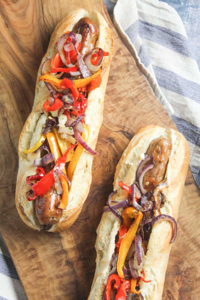 Two sandwich baguettes filled with cooked pork sausages and roast sliced peppers and onions. 