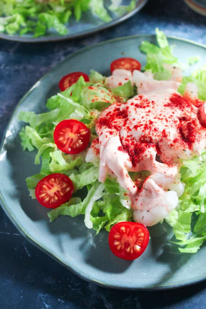 Close up shot of prawn cocktail. There is a blue plate topped with shredded romaine lettuce, sliced cherry tomatoes and prawns. The prawns are garnished with a pink cocktail sauce and sprinkled with paprika. 