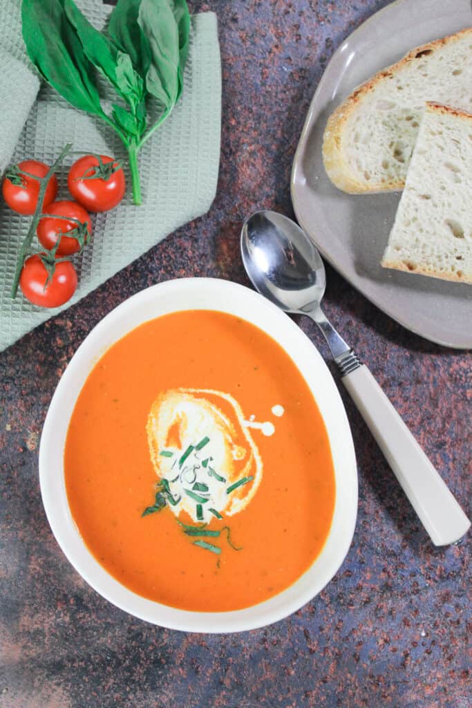 White bowl with tomato and red pepper soup next to a small plate with slices of bread on it.