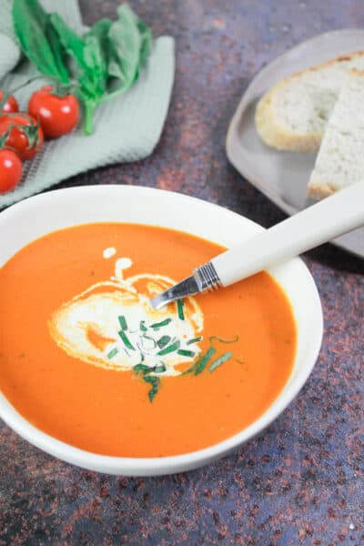 White bowl filled with tomato and red pepper soup. The soup is garnished with a drizzle of cream and chopped basil.