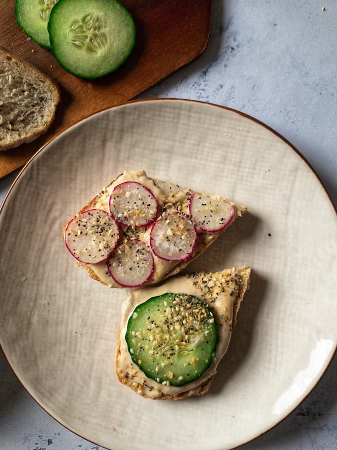 Slices of sourdough toast spread with hummus and topped with sliced cucumber and radishes with a sprinkle of seeds. 