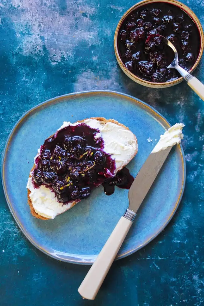Slice of sourdough toast spread with white ricotta cheese and topped with blueberry compote.