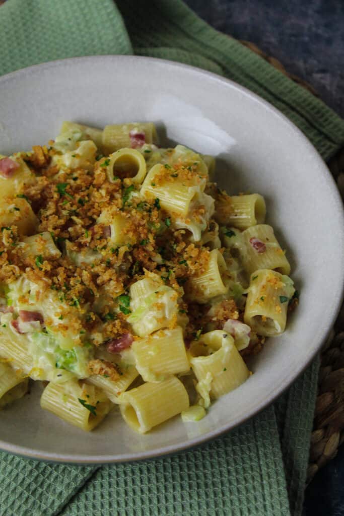White pasta bowl filled with rigatoni pasta in a cream sauce with sliced leeks and pancetta cubes. The pasta is topped with golden breadcrumbs and chopped parsley.