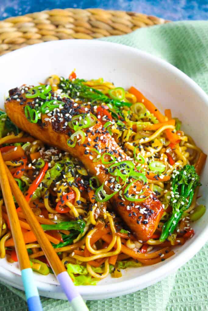 Salmon fillet on a bed of noodles tossed with sliced pepper, broccoli and carrot. The salmon is glazed with a dark orange teriyaki sauce and garnished with sliced spring onion and sesame seeds.