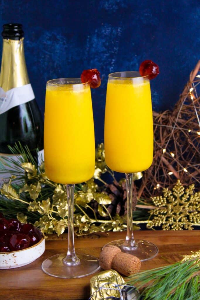 Two champagne flutes filled with fresh orange juice and non-alcoholic sparkling wine and garnished with a glace cherry. 