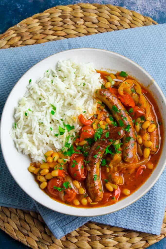 Sausage and bean casserole in a tomato sauce with sliced peppers, served on a bowl of white rice. 