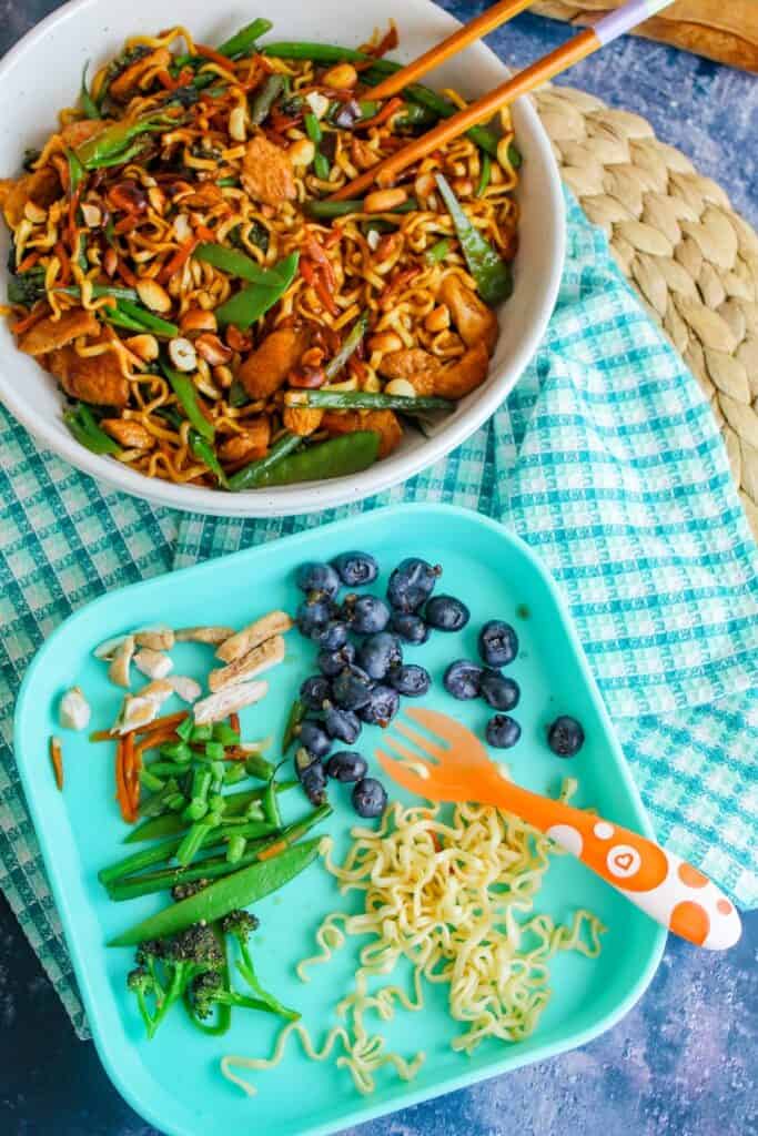 Stir-fried egg noodles with vegetables and chicken in a white bowl on a blue-checkered cloth.