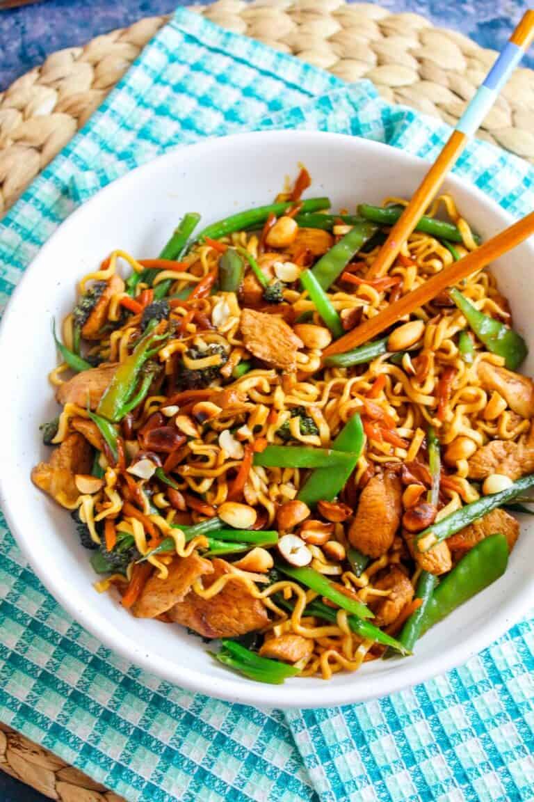 Stir-fried Asian noodle dish with vegetables, chicken, and peanuts in a white bowl on a blue checkered cloth.